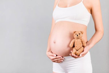 Elinde oyuncak ayının kesilmiş görüntüsü gri arka planda beyaz iç çamaşırlı hamile kadının göbeğine karşı. Çocuk doğurmayı bekliyor. Boşluğu kopyala.