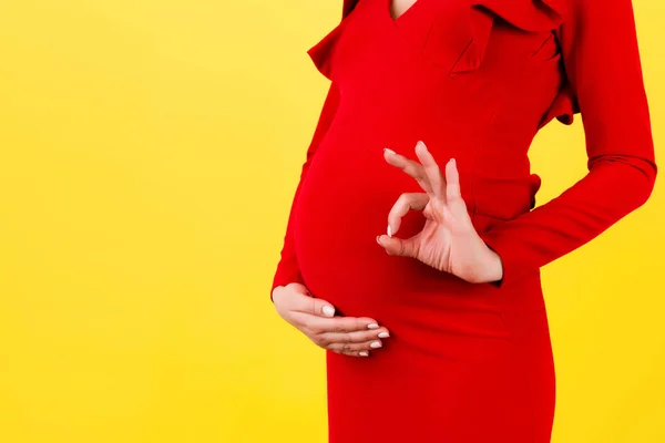黄色の背景で彼女の腹部の上に大丈夫なジェスチャーを示す赤いドレスの幸せな妊婦のクローズアップ 肯定的な妊娠 スペースのコピー — ストック写真