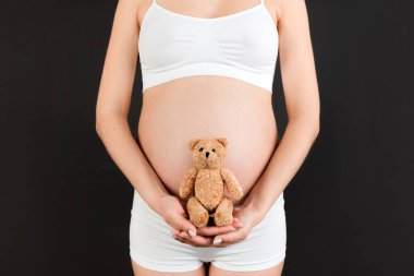 Beyaz iç çamaşırlı hamile kadının siyah arka planda karnına oyuncak ayıyı dayadığı yakın plan. Ebeveynlik kavramı. Boşluğu kopyala.