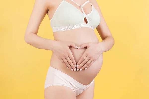 用黄色背景的手指做的心脏紧闭孕妇的腹部 产妇概念 爱的未来的宝宝 复制空间 — 图库照片