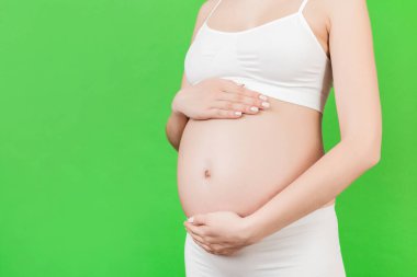 Yeşil arka planda karnını kucaklayan hamile bir kadının ellerinin kesilmiş görüntüsü. Gelecekteki anne beyaz iç çamaşırı giyiyor. Bir bebek bekliyorduk. Boşluğu kopyala.