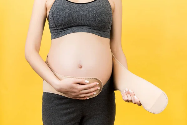 Cropped image of pregnant woman on the third trimester putting on pregnancy belt at yellow background. Orthopedic abdominal support belt concept with copy space.