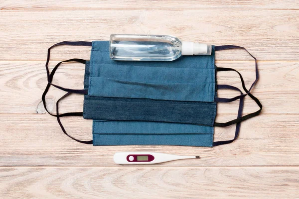 Top view of handmade cotton masks, digital thermometer and alcohol gel sanitizer on wooden background. Protective equipment concept with copy space.