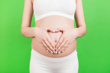 Beyaz iç çamaşırlı hamile bir kadının yeşil arka planda elleri karnında kalp şekli oluşturduğu yakın plan. Gelecekteki bebeğe sevgiler. Boşluğu kopyala.