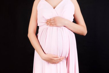 Siyah arka planda karnını kucaklayan hamile bir kadının ellerinin kesilmiş görüntüsü. Müstakbel annem pembe elbise giyiyor. Bir bebek bekliyorduk. Boşluğu kopyala.