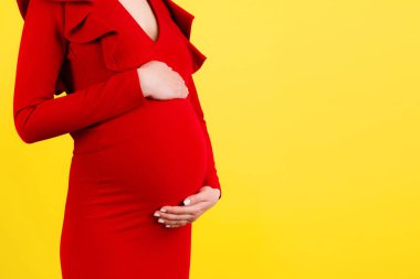 Kırmızı elbiseli hamile bir kadının sarı arka planda koca karnını kucaklarken görüntüsü. Annelik konsepti. Boşluğu kopyala.