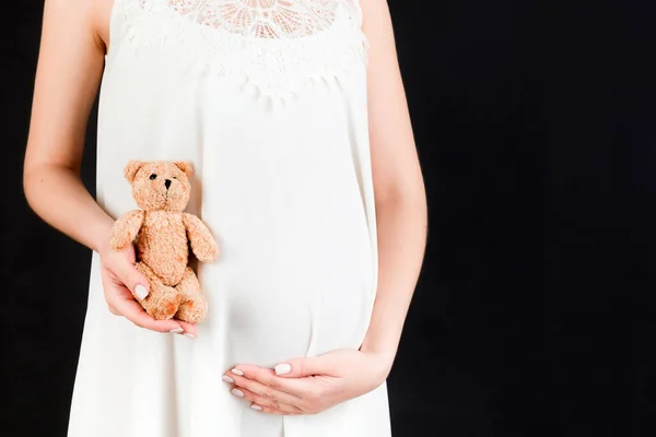 黒を背景に白いドレスを着た妊婦のお腹に手をつないでテディベアの姿を切り取った 出産を待っている スペースのコピー — ストック写真