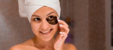 Beyaz havluya sarılı gülümseyen kadın, sabah duş aldıktan sonra mor göz bandı yapıştırıyor. Gözlerinin altındaki siyah halkaları çıkar..