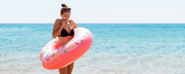 Sıska kadın elinde donut şişirilebilir yüzüğüyle suda duruyor ve üşüyor. Deniz arkaplanı.