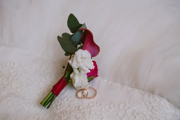 Close-up de anéis dourados e boutonniere no fundo de um vestido de noiva de renda. Preparação para o conceito de casamento . — Fotografia de Stock