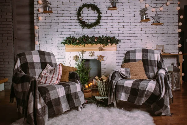 Dwa fotele pokryte przytulnymi kocami stojącymi przy kominku ozdobionym girlandą, świecami i koszykiem z prezentami. Okrągły zielony wieniec wiszący na białej ceglanej ścianie. Świąteczne wnętrze. — Zdjęcie stockowe