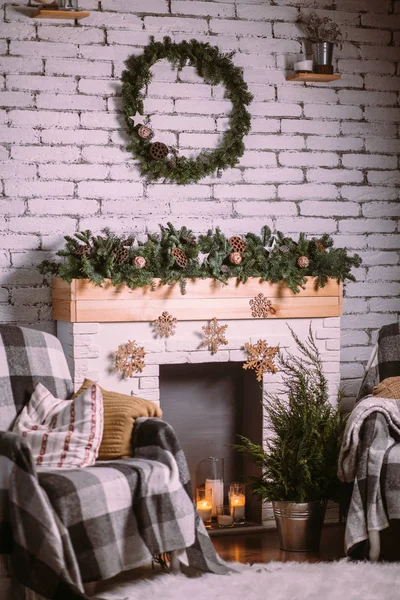 Διακοσμητικό τζάκι διακοσμημένο με γιρλάντα και αναμμένα κεριά σε γυάλινες κούπες. Στρογγυλό πράσινο στεφάνι που κρέμεται σε λευκό τοίχο από τούβλα. Δροσερή ατμόσφαιρα Χριστουγέννων. Royalty Free Εικόνες Αρχείου