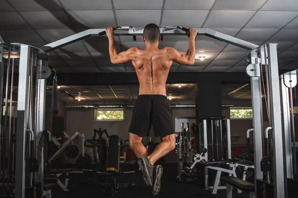 Een jonge atleet traint zijn rugspieren op een horizontale balk met een brede grip. Lessen in de sportschool, gezonde levensstijl. — Stockfoto