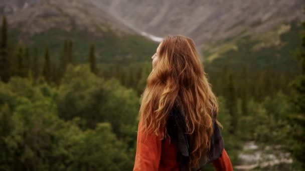 Молода мандрівниця з довгим волоссям стоїть з піднятими руками на вершину, захоплюючи гірський ландшафт. — стокове відео