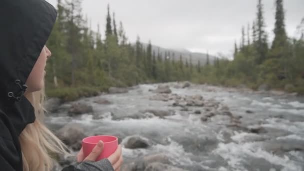 Close up mulher viajante relaxar perto de um riacho de montanha e desfrutar de chá quente ou café em uma caneca . — Vídeo de Stock