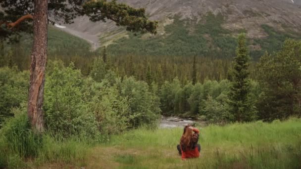 Junge Wanderin mit langen Haaren sitzt im Gras und blickt in die Berge — Stockvideo