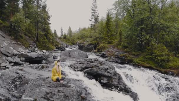 Sarı yağmurluklu genç kadın dağlardaki şelalenin yanında yoga yapıyor.. — Stok video