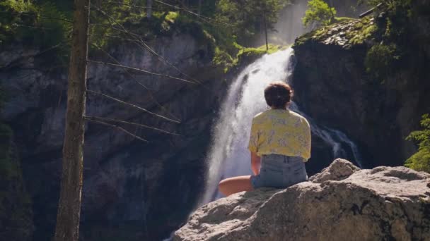 裏面ビュー若い女性が大きな岩の上に座って崖からの滝のストリーミングを見て スローモーション — ストック動画