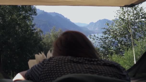 湖や山を背景に美しい本を読みながらテントの中で寝そべっている若い女性 — ストック動画