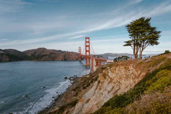 Ünlü Golden Gate Köprüsü 'nün yatay fotoğrafı. Okyanusun üzerindeki kayalık uçurum ve köprünün arkasındaki yeşil ağaç.