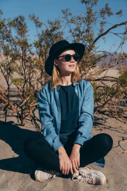 Kaliforniya 'daki Ölüm Vadisi Ulusal Parkı' nda, geniş kenarlı siyah bir ağacın yanında oturan bir kız keçeli şapka takıyor. Ölüm Vadisi çölünde poz veren mavi gömlekli bir gezgin kız.