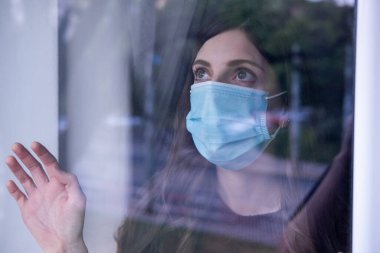Coronavirus için tıbbi maske takan ve pencereden dışarı bakan genç bir kadın. Covid 19.