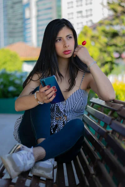 Kolumbianerin checkt ihr Handy, sitzt in einem Holzstuhl und isst ein rotes Bonbon — Stockfoto