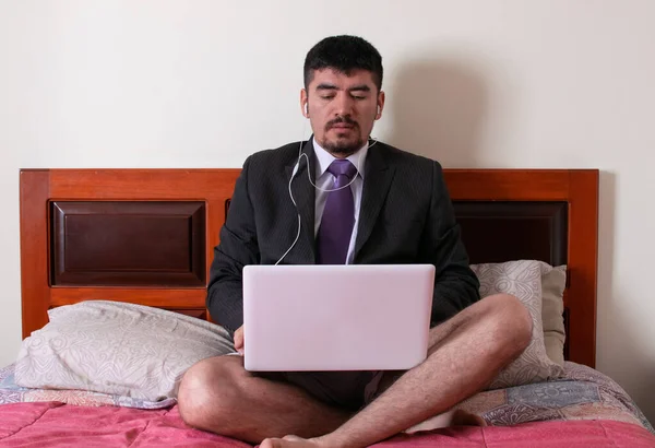 man latin american in formal suit sitting on his bed with headphones on is in a business meeting at his laptop