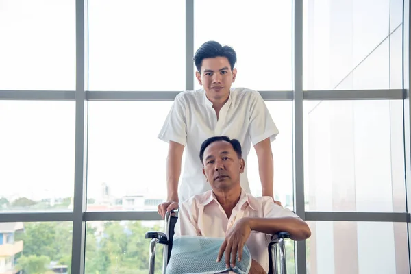 Elderly Asian man has an arm injury sitting in a wheelchair in a hospital with a young son, a happy caretaker, the concept of caring for the elderly.