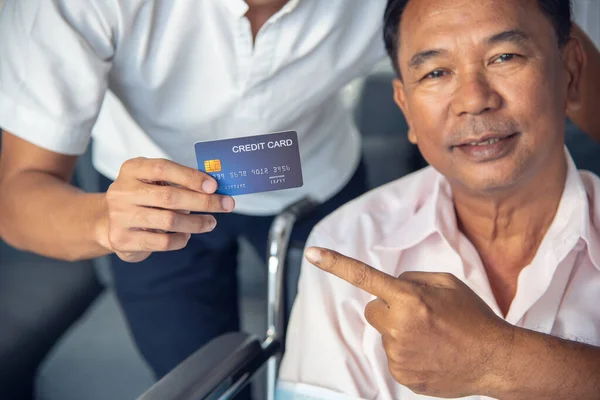 Seior Asian man has an arm injury sitting in a wheelchair, using a special promotion card to pay hospital bills with a young son, a happy caretaker, the concept of caring for the elderly.