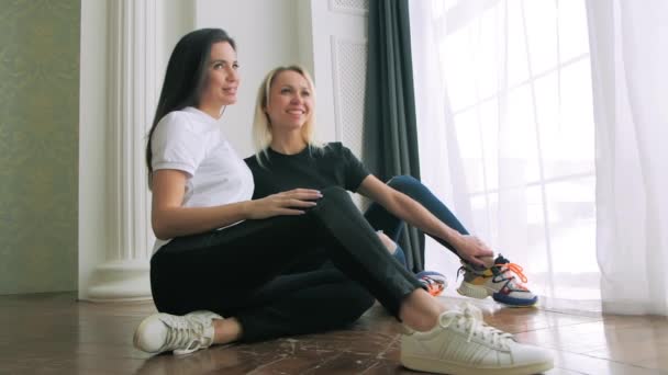 Két tornacipős lábú nő ül a földön. Szexi lány barátok mosoly közelkép. — Stock videók