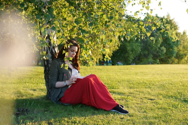 Jovem mulher senta-se com um livro sob uma árvore em um dia ensolarado — Fotografia de Stock