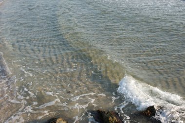 Güneşli bir gün, deniz berrak su ve kumsalda dalgalar var.