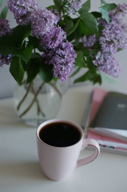 Cam vazolu bir fincan çay, mor leylak ve masa arkasında kitaplar olan bir laptop.