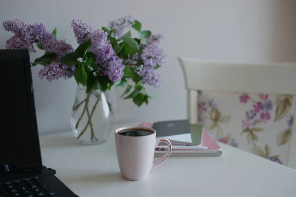 cup of tea with glass vase with purple lilac and laptop with books on table background