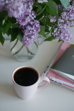 Cam vazolu bir fincan çay, mor leylak ve masa arkasında kitaplar olan bir laptop.