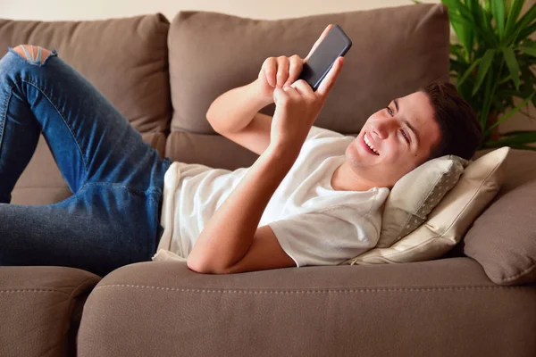 Fiatal fiú mosolygó arc fel a kanapén egy smartphone tanácsadás — Stock Fotó