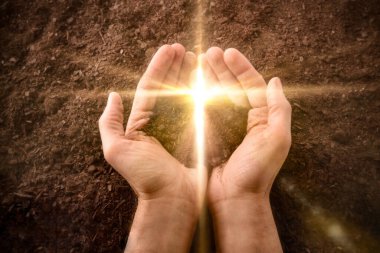 Hands together with a small mound of earth with a golden flash in the shape of a cross on a background of earth. Jesus resurrection concept. Horizontal composition. Top view. clipart