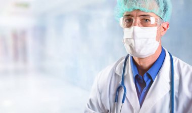 40 'lı yaşlardaki doktor maske, gözlük ve şapka takıyor. Hastane koridorunda boynunda steteskop asılı beyaz bir üniforma giyiyor.