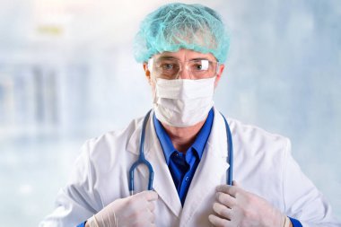 40 'lı yaşlardaki doktor maske, gözlük ve şapka takıyor. Hastane koridorunda boynunda steteskop asılı beyaz bir üniforma giyiyor.