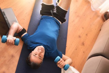 Oturma odasında halterli spor yapan bir adam, kulaklık ve telefonla müzik dinliyor. Üst görünüm