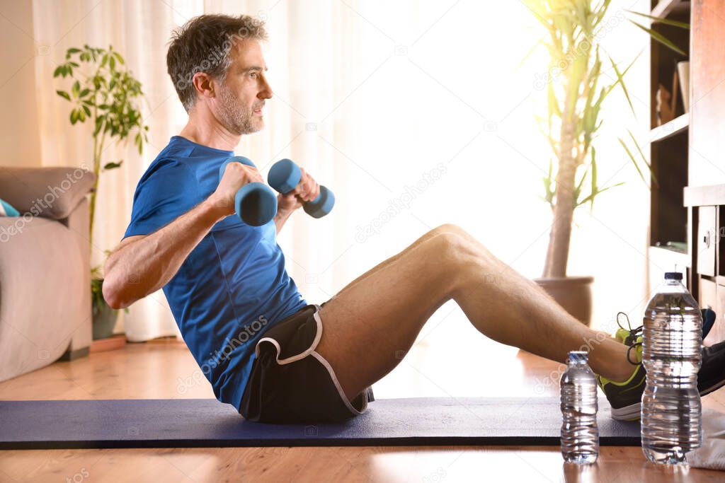 Man doing dumbbell exercises sitting on mat in his living room watching a television