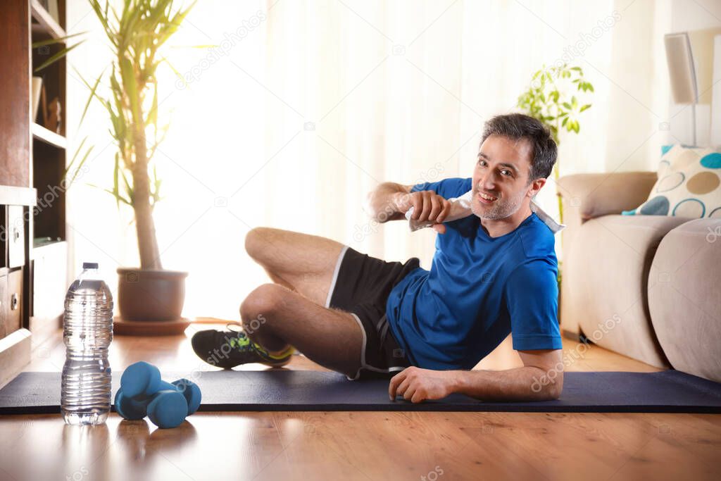 Man prepared to do sports at home lying on mat smiling looking straight ahead with sports equipment on the floor in the living room at home