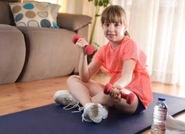 Komik küçük kız mutlu gülümsüyor ve halterlerle spor yapıyor. Evdeki oturma odasında bir paspasın üzerinde oturuyor.