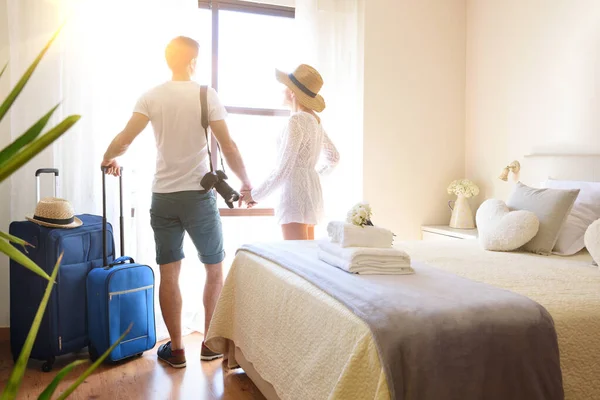 Parlak bir otel odasında el ele tutuşup pencereden dışarı bakan çift tatilde..