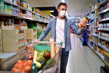 Maskeli ve eldivenli bir adam alışveriş arabasını itiyor ve süpermarkette gıda ürünleri alıyor.