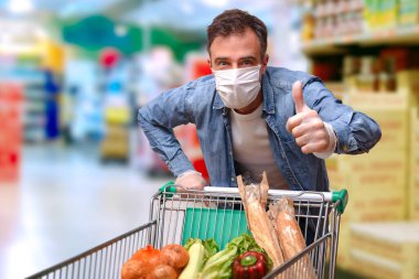 Maskeli ve eli eldivenli koruma altında bir süpermarketten yiyecek dolu bir araba almak.