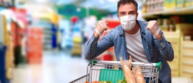Maskeli, eldivenli, yumrukları kapalı, başarılı bir jest yapan, içi yiyecek dolu bir arabayla süpermarketten alışveriş yapan koruma altında bir adam.