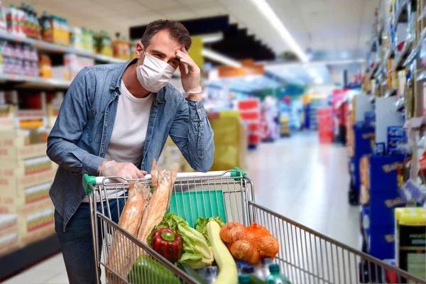 Sick man with fever leaning on a shopping cart full of food with face shields and gloves in a supermarket