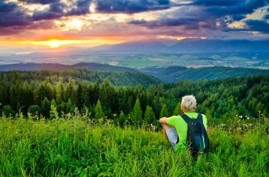 Genç adam güzel yeşil çimenlerde oturuyor ve çarpıcı bir şekilde karanlık gökyüzünde renkli günbatımını seyrediyor. Yeşil ağaçlar ve çayırlarla dolu güzel manzaranın üzerine güneş düşüyor.